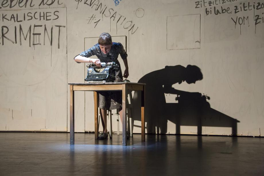 Frau beugt sich von hinten über eine Schreibmaschine, die vor ihr auf dem Tisch steht.