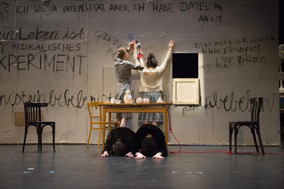 Eine beschriftete Wand, davor ein Tisch, dahinter zwei Frauen. Eine Frau schlägt mit dem Hammer einen Nagel durch die Hand der anderen Frau. Vor dem Tisch knien in Gebetshaltung zwei schwarz gekleidete Menschen, deren Gesichter verborgen sind.