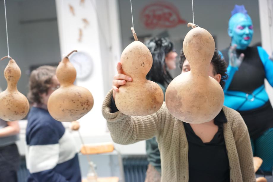 4 Kürbisse hängen an Schnüren an der Wand. Im Hintergrund seteht ein Mensch mit blauer Haut und Irokesenschnitt.