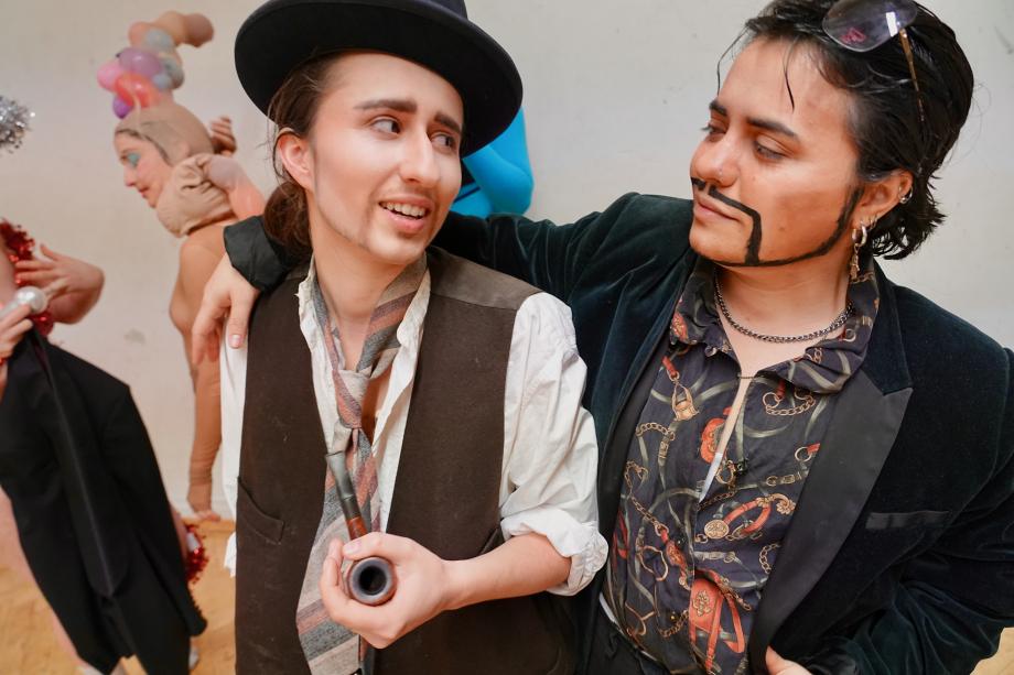 zwei Frauen als Männer verkleidet mit aufgeschminktem Bart und Pfeife in der Hand umarmen sich und schauen sich dabei an.