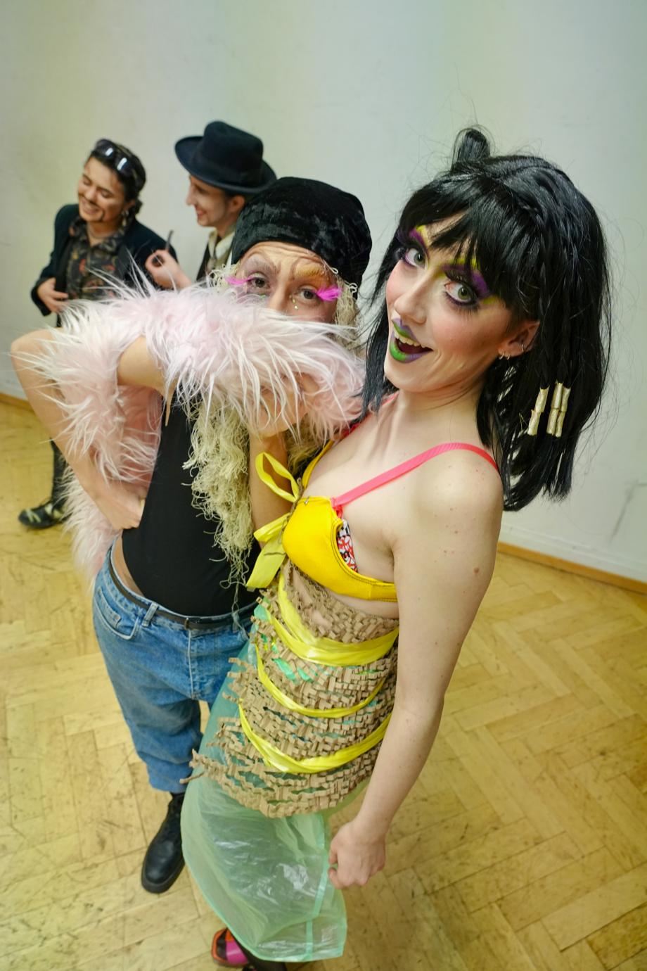zwei bunt geschminkte und bunt angezogene Frauen schauen in die Kamera. Im Hintergrund sind zwei Menschen zu erkennen.