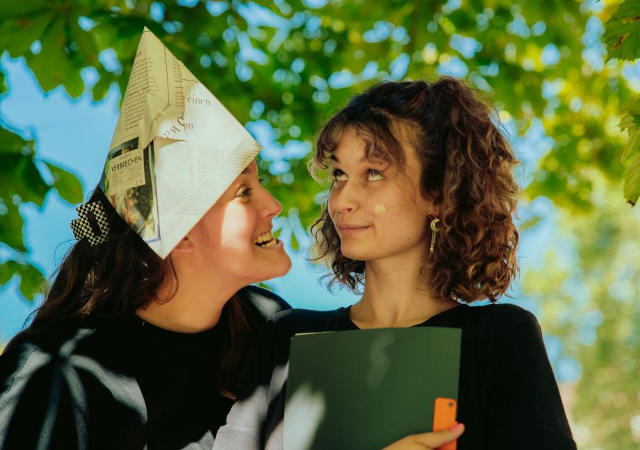 Frau mit Papierhut schneidet eine Grimasse und schaut dabei eine andere Frau an. Diese hält eien Mappe und verdreht die Augen.