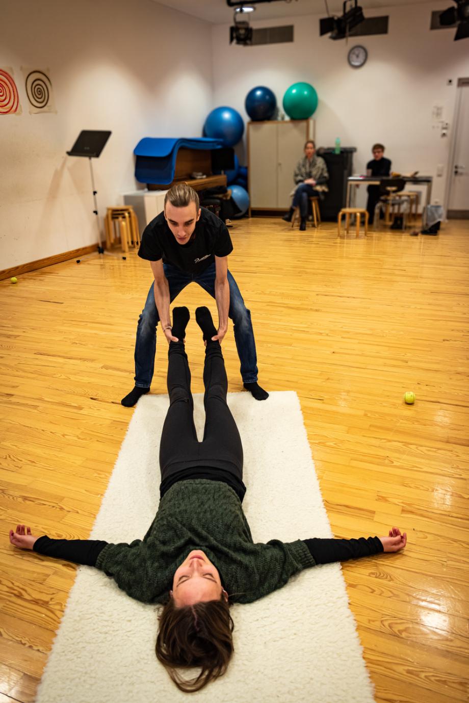 Im Rahmen der Bewegungsarbeit hebt ein Studient die Beine einer Studentin, die auf einer weißen Matte liegt, kniehoch an. Im Hintergrund beobachten zwei Studierende die Situation.