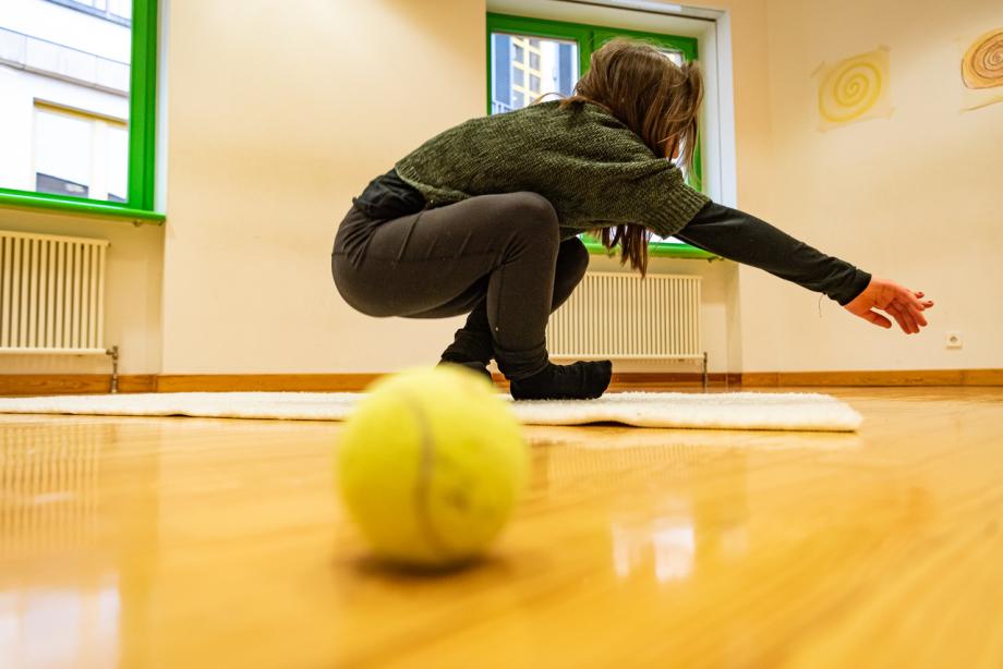 Tennisball im Vordergrund. Dahinter Studentin, die sich auf eine Matte setzt.
