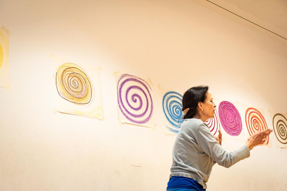 Frau gestikuliert. Hinter ihr an der Wand farbige gemalte Kreise.