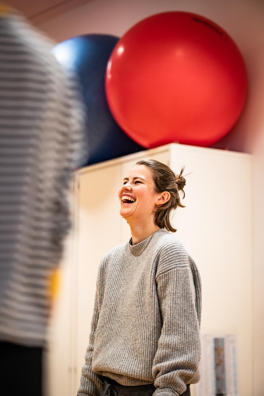 Lachende Studierende während einer Bewegungsarbeit. Im Hintergrund sind Medizinbälle zu sehen, die aus der Beobachtungsperspektive, scheinbar auf ihrem Kopf balancieren.