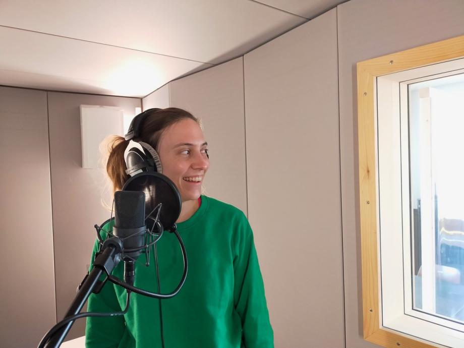 Studentin lacht mit Kopfhörern vor Mikrophon in Sprechkabine.