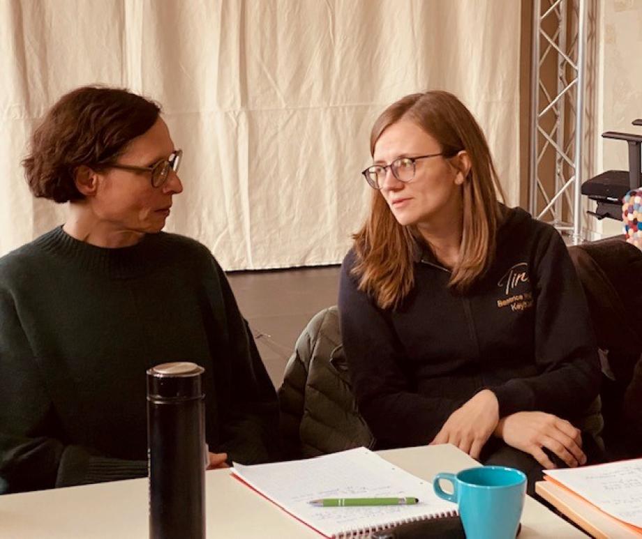Zwei Frauen, eine mit kurzen, Ohrlnagen dunkel braunen Haaren, die andere mit dunkelblonden Schulterlangen offenen Haaren sitzen nebeneinander an einem Tisch und sprechen miteinander.