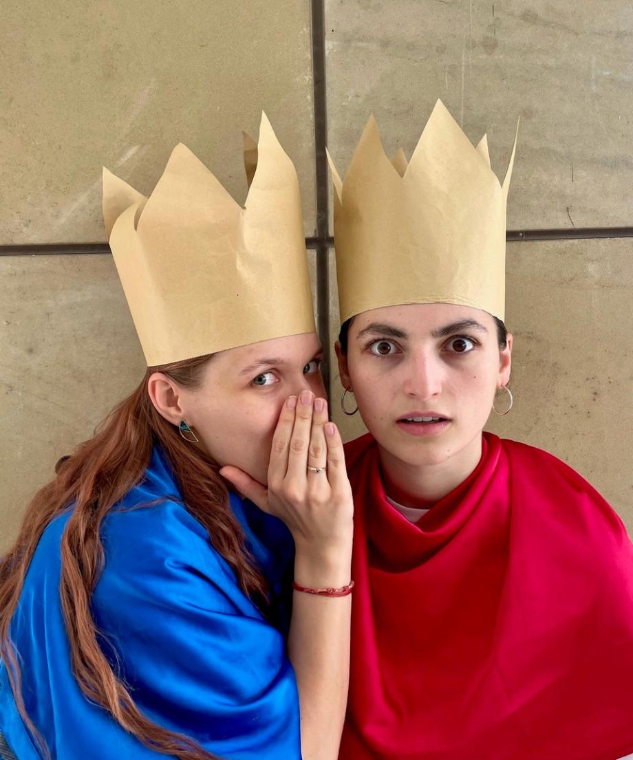 Eine Studierende mit Krone flüstert einer anderen bekronten Studierenden etwas zu. 