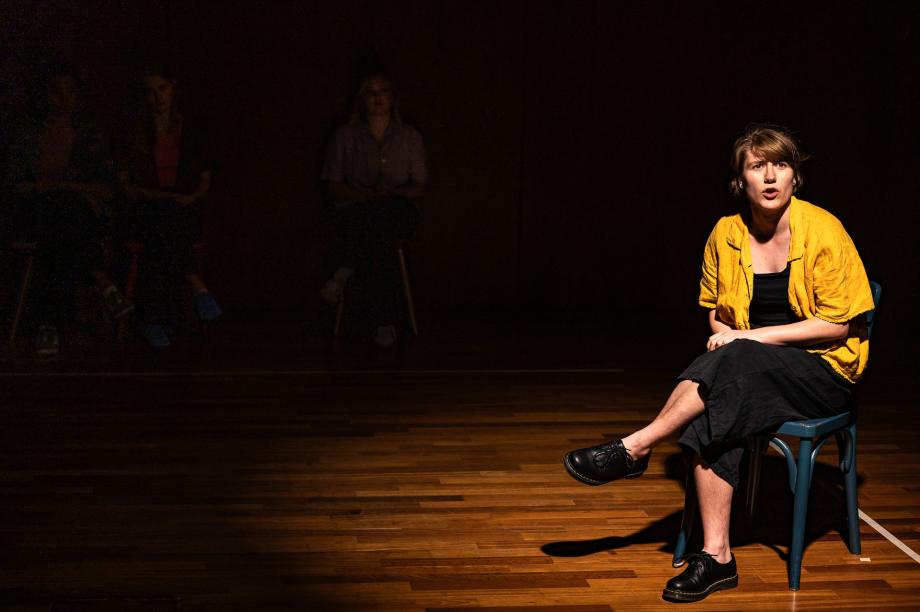 Studierende sitzt am Bildrand auf einem Stuhl, die Beine übereinander geschlagen und spricht ins Publikum. Der Raum um sie herum ist dunkel. Ein Lichtspot scheint auf sie.