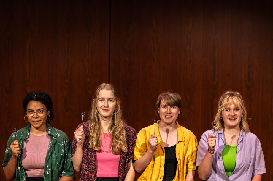 Vier Studierende neben einander in farbenfrohen Kostümen. Jede hällt einen Löffel in der Hand und spricht lächelnd ins Publikum.