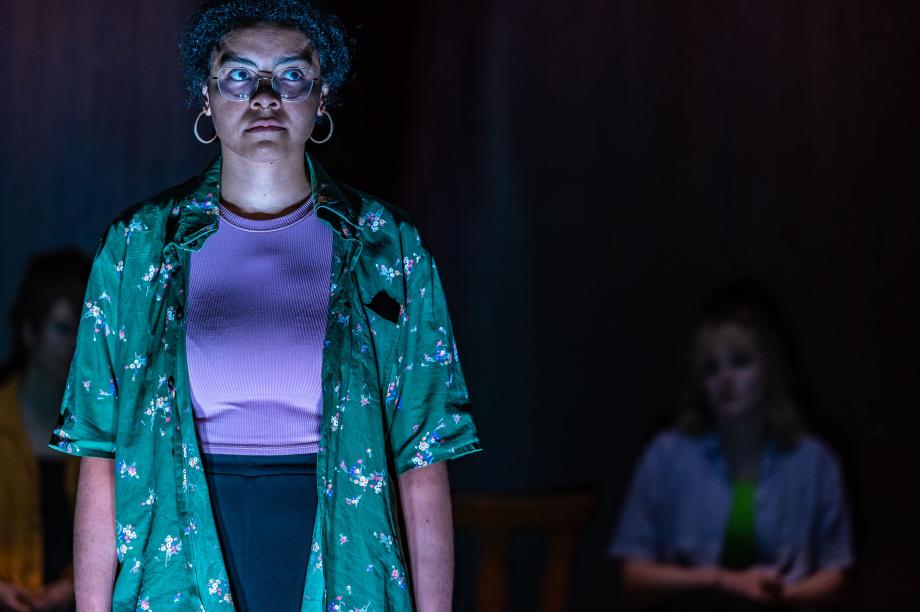 Studierende steht frontal zum Publikum. Ihr Mund ist geschlossen und sie blickt seitlich nach oben. Im Hintergrund verschwommen eine weitere Studierende.