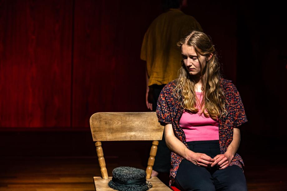 Studierende sitzt auf einem Stuhl. Neben ihr ein weiterer Stuhl, auf dem ein Hut liegt. Sie betrachtet diesen still.