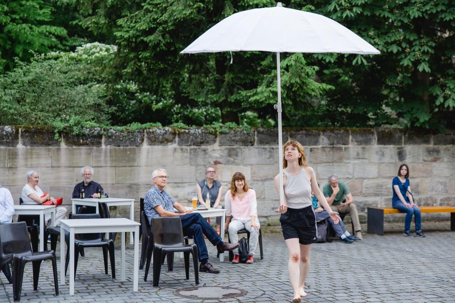 Frau balanciert Sonnenschirm auf ihrer Hand vor einem Publikum an Tischen, das hinter ihr sitzt.
