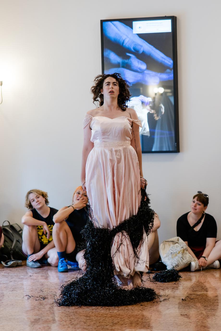 Eine Frau in einem langem rosa Kleid steht in schwarzen Kunststofffäden und schaut an die Decke. Hinter ihr sitzen Menschen, die die Frau anschauen.