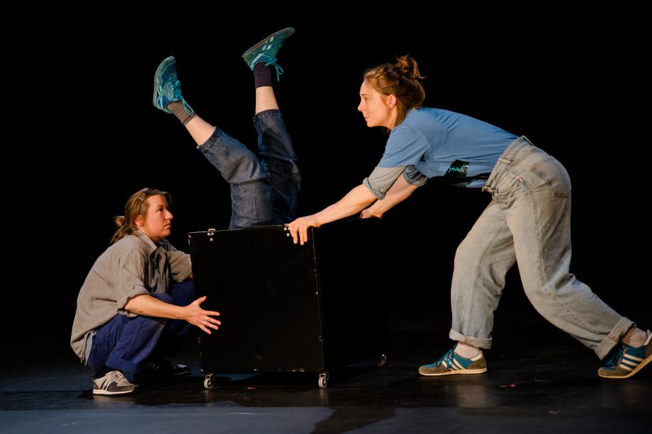 zwei Frauen schieben einen Kasten auf Rollen. Aus dem Kasten schauen zwei Beine.