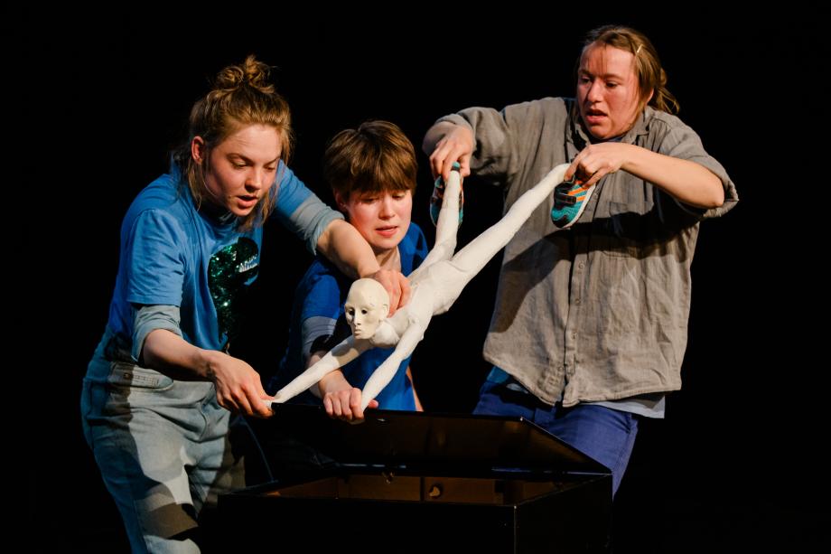 Drei Frauen halten die Figur in der Luft. Eine Frau hält und streckt die Arme der Figur, die andere Frau hält und zieht an den Beinen.