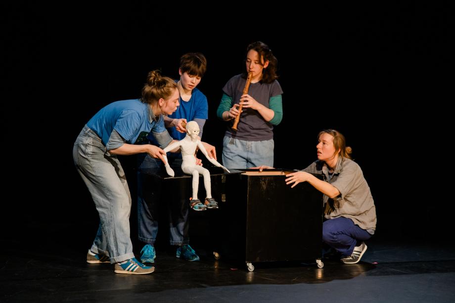 vier Frauen interagieren mit einer kleinen sitzenden Figur. Eine Frau spielt Flöte.