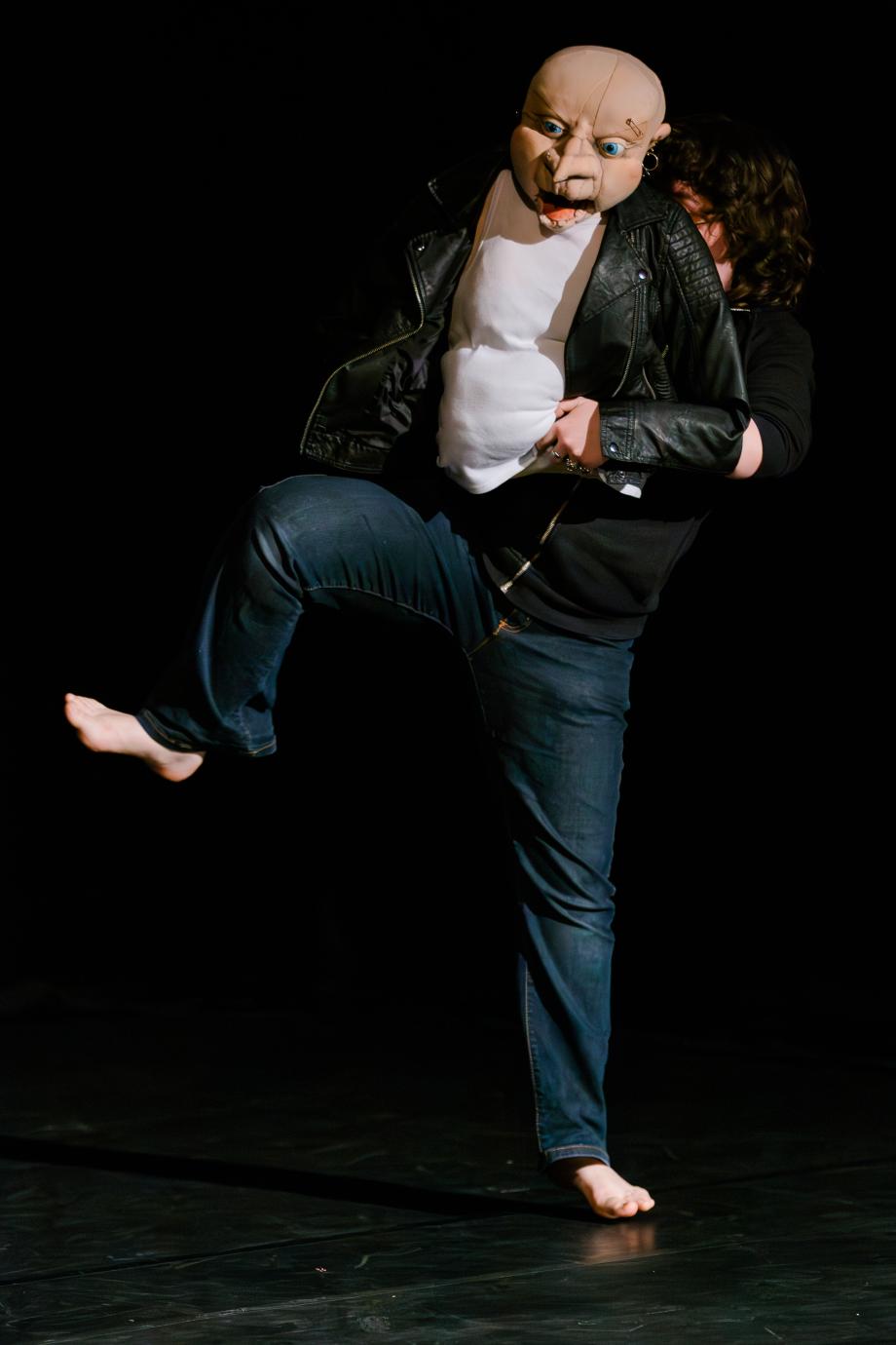 Figur mit Glatze und gepircten Augenbrauen und Ohrringen wird von einer Figurenspielerin im Hintergrund geführt.