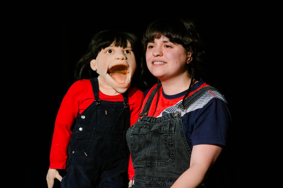 Frau hält eine relativ große Figur neben sich in der Hand. Die Frau und die Figur tragen ähnliche Kleidung und eine ähnliche Frisur.