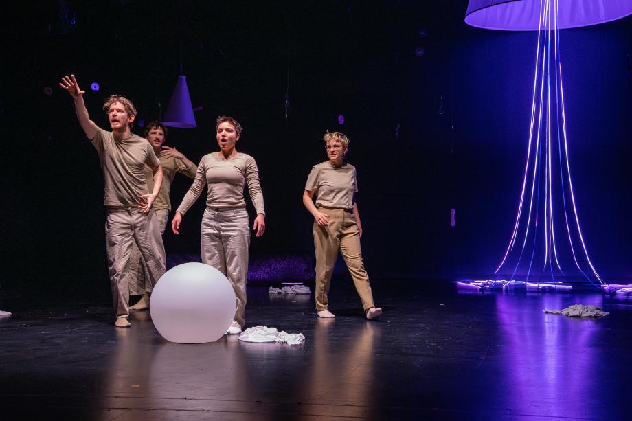 Vier Studierende mit beigfarbenen Kostümen auf der Bühne. Einer streckt die Hand ins Publikum und spricht.