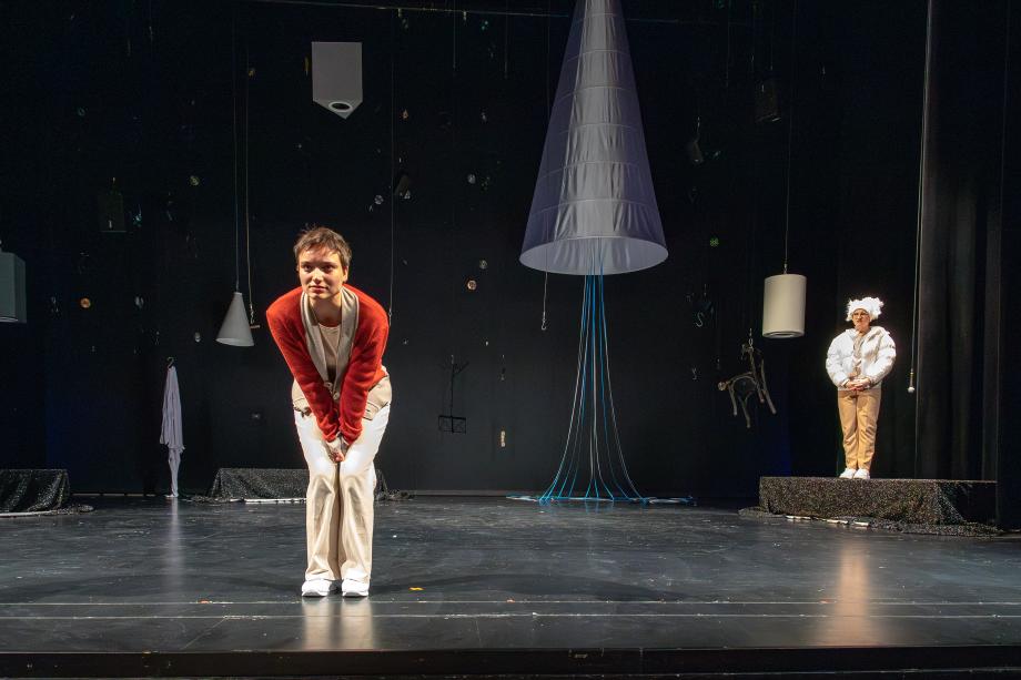 Studentin steht nach vorne zum Publikum gebeugt auf der Bühne, die Hände zwischen die Knie geklemmt. Im Hintergrund eine weitere Person im Kostüm mit weißem Puschelhut.