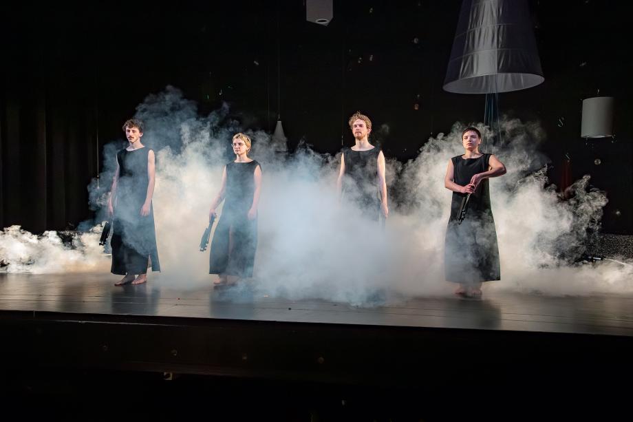 Vier Studierende in langen schwarzen Kostümen stehen in einer Reihe auf der Bühne und schauen ins Publikum. Um sie herum beleuchteter Theaternebel.