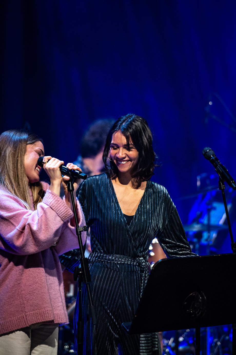 Zwei junge Jazzsängerinnen lachen sich auf der Bühne an.