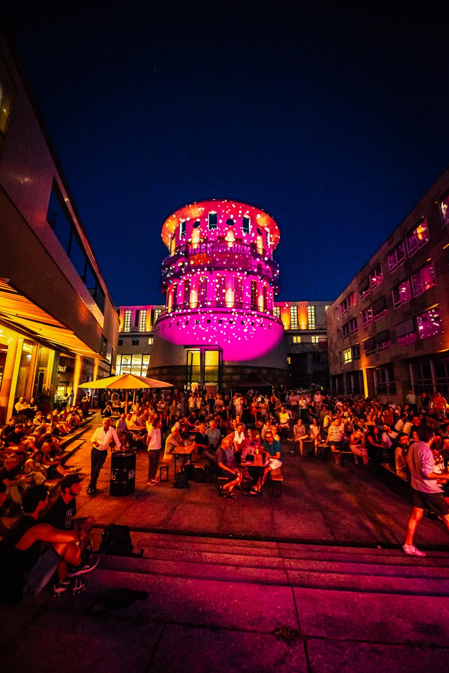 Die Piazza und der Turm der Musikhochschule sind bunt beleuchtet. Bei der Nachtstimmung ist der Platz voller Menschen.