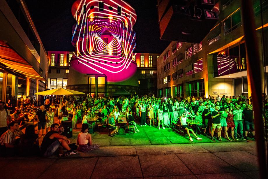 Stehende und sitzende Zuhörer*innen verfolgen ein Konzert auf der Bühne im Freien. Alles ist bunt beleuchtet.