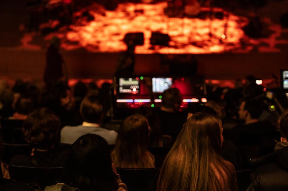 Zuschauer*innen im dunklen Konzertsaal schauen auf die beleuchtete Bühne und warten auf den Konzertbeginn.
