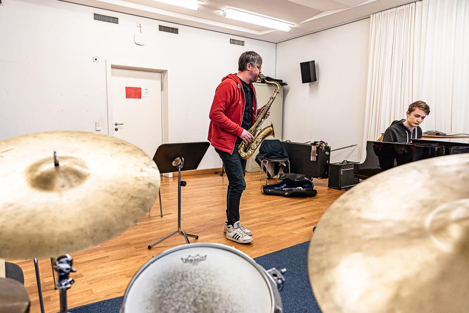 Im Unterricht spielt ein Dozent Saxophon mit einem Studierenden, der Klavier spielt. Im Raum steht auch ein Schlagzeug.
