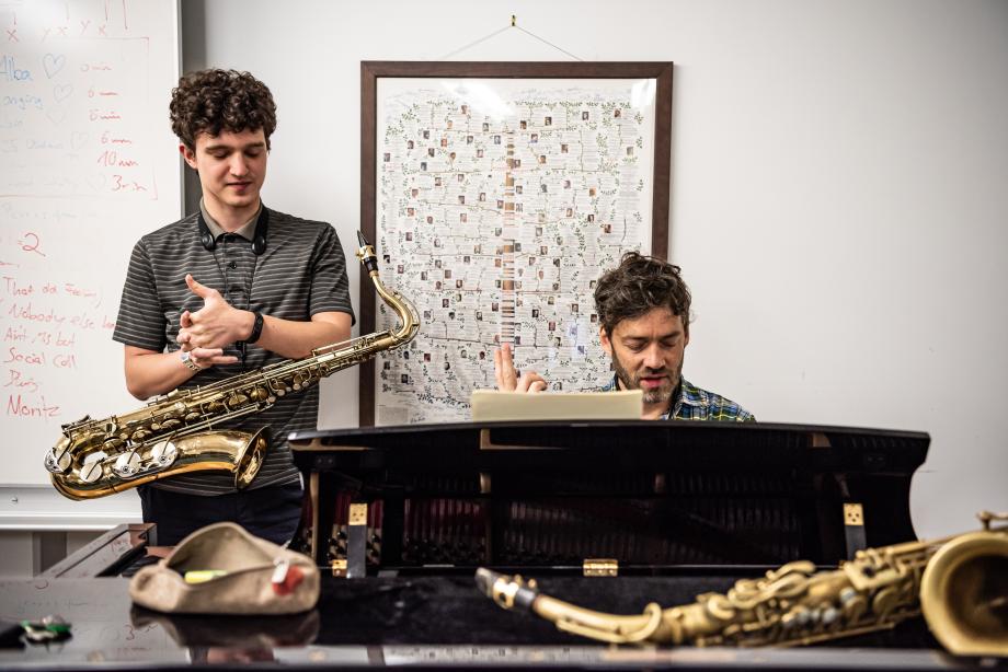 Im Saxophonunterricht spricht der Lehrer mit seinem Studenten. Sie schauen aufs Klavier, an dem der Lehrer sitzt.
