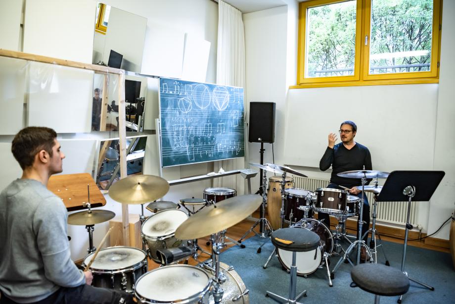 Ein Schlagzeug-Student sitzt seinem Lehrer gegenüber, beide an einem eigenen Schlagzeug. Daneben steht eine Kreidetafel mit rhythmischen Notizen.