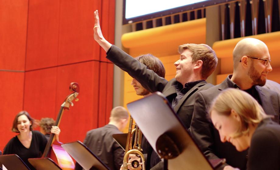Ein Musiker auf der Bühne winkt einer Person im Publikum vor Konzertbeginn. Die andern Musiker*innen lachen.