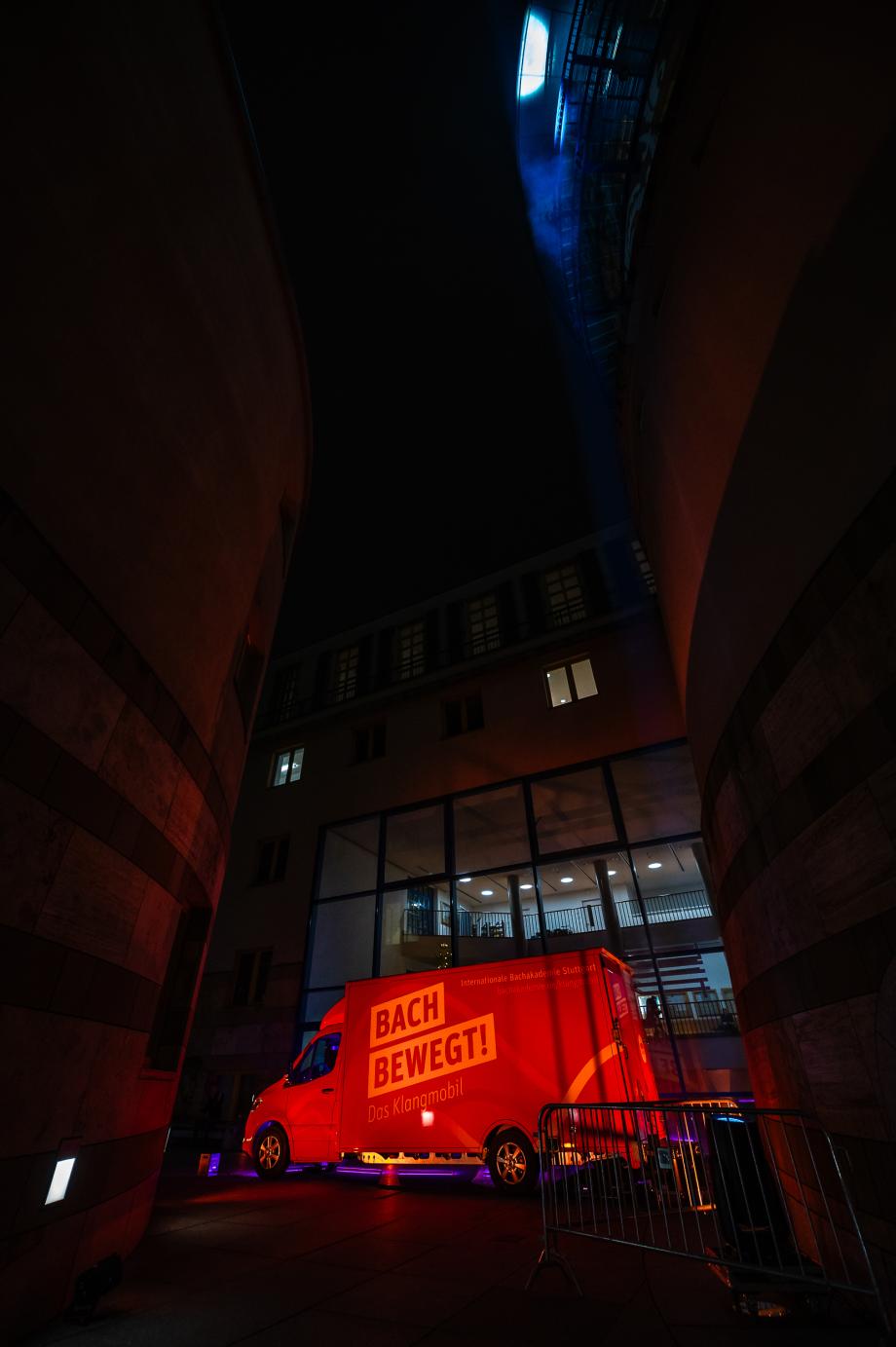 Das Klangmobil unter bunter Beleuchtung nachts, im Hof der Musikhochschule stehend. Im Hintergrund kann man durch Glasfenster in die Hochschule hineinsehen.