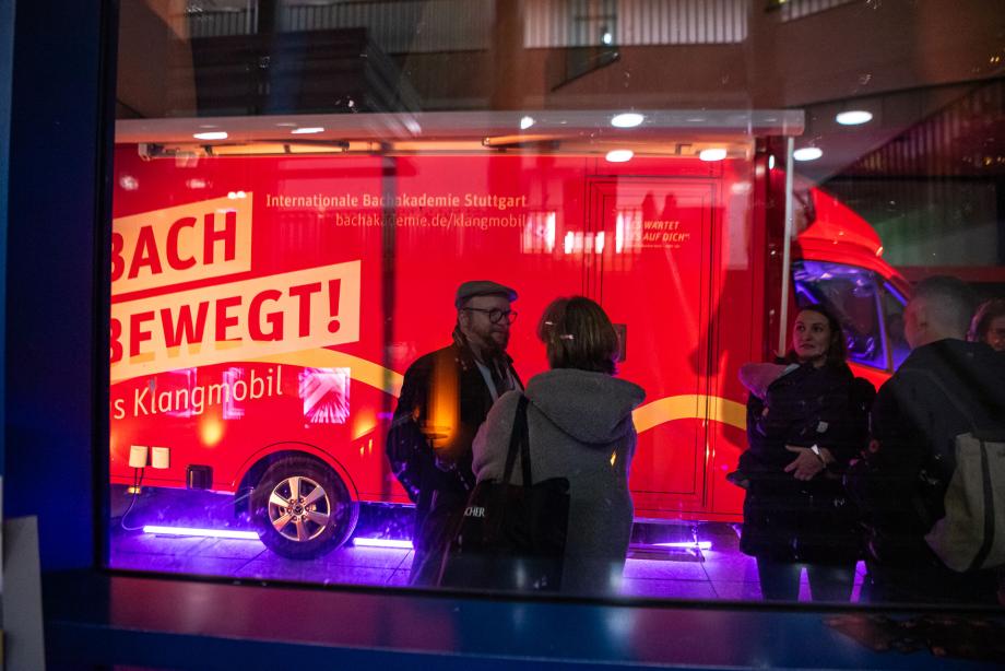 Rotes Fahrzeug mit der Aufschrift 'BACH BEWEGT! Das Klangmobil' und mehreren Personen davor in einer nächtlichen Szene