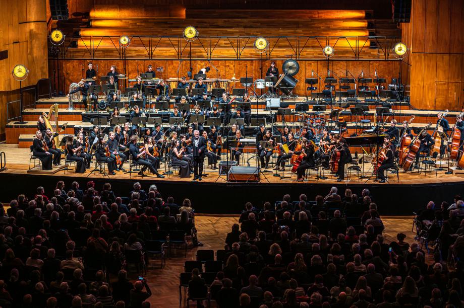 Blick von der Zuschauerempore im ersten Rang auf die Bühne des Beethovensaales. Axel Köhler, Rektor der HMDK Stuttgart, begrüßt das Publikum. Auf dem Podium sitzt das Hochschulsinfonieorchester mit rund 80 Studierenden.