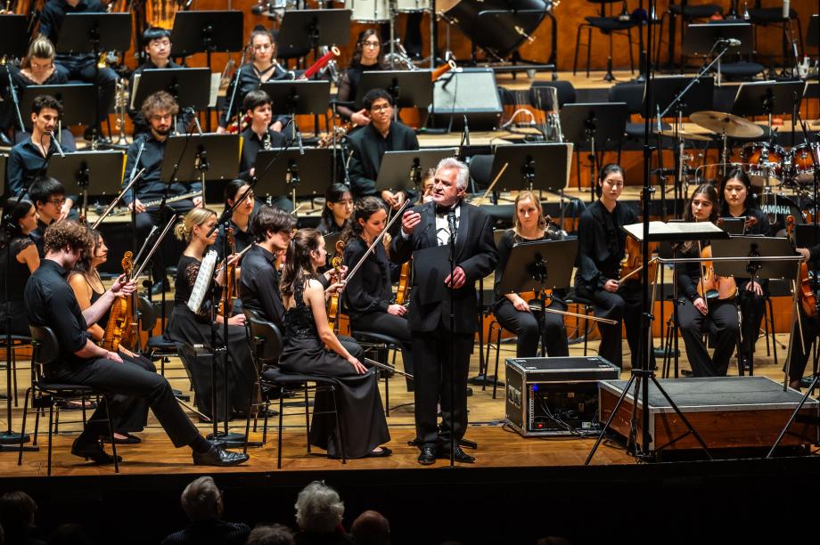 Axel Köhler bei der Begrüßung zum Tributekonzert für Wolfgang Dauner. Im Hintergrund sitzen die Musikerinnen und Musiker des Hocshchulsinfonieorchesters.