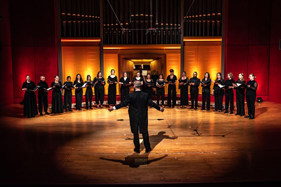Der Kammerchor der Hocschule ist im Halbkreis auf dem Konzertsaalpodium positioniert. Im Vordergrund ist mit dem Rücken zum Publikum zugewandt der Chorleitungsprofessor Denis Rouger zu sehen. angezogen. Vor ihnen ein Dirigent.