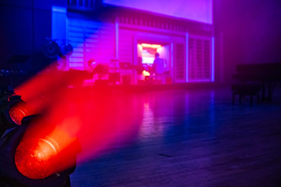 Roter Scheinwerfer, der eine Bühne beleuchtet. Im Hintergrund sieht man jemanden schemenhaft an einer Orgel sitzen.