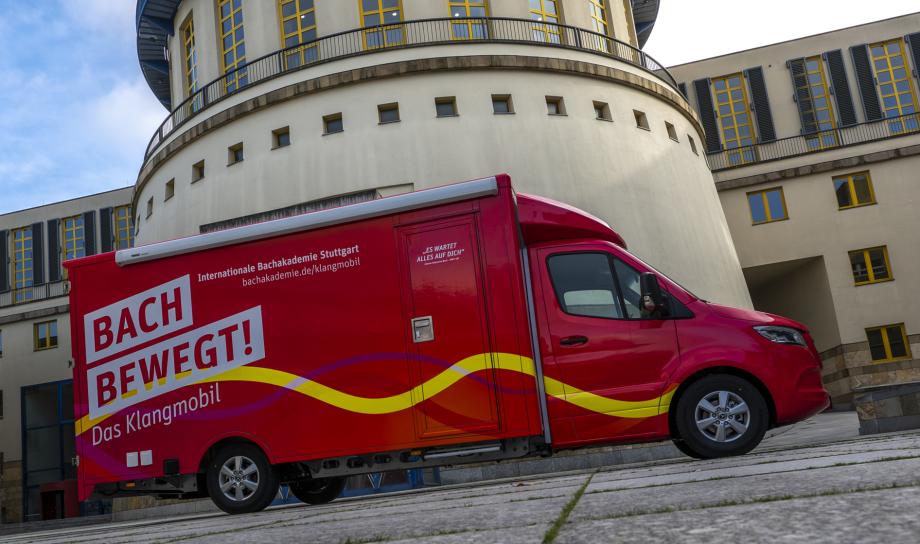 Ein roter Transporter mit der Aufschrift „Bach bewegt!“ und farbigen geschwungenen Linien auf der Seite