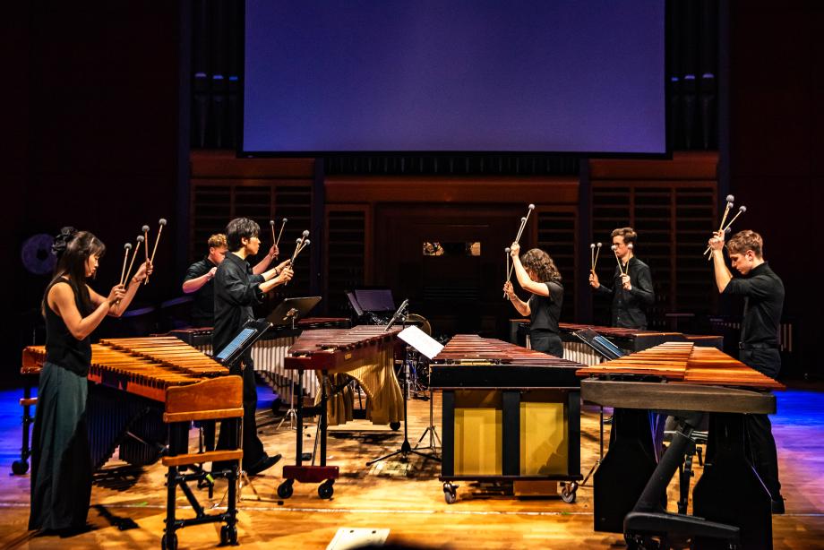 Studierende der Schlagzeugklasse musizieren mit sechs Mibraphonen. Die Schlaginstrumente sind so angeordnet, dass zwei Mibraphone sich in der Mitte verbinden und von den vier weiteren Instrumenten umgeben werden. Alle Mitwirkenden haben die Hände mit jeweils zwei Schlägeln in die Luft gehoben.