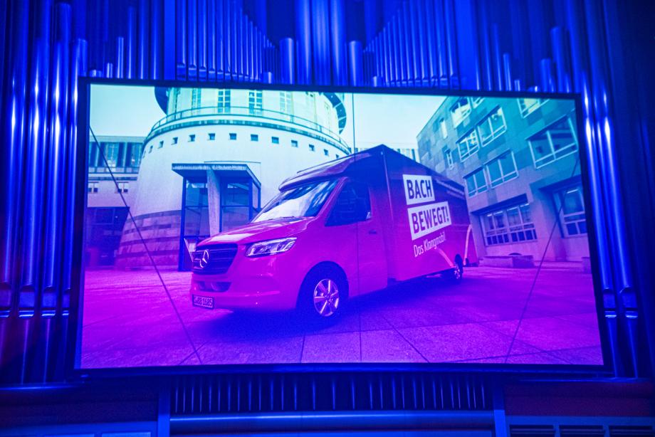 Vor großen Orgelpfeifen ist ein Bildschirm zu sehen. Darauf abgebildet sieht man das Klangmobil, ein roter Mercedeslieferwagen, mit der Aufschrift 'BACH BEWEGT! Das Klangmobil' vor dem Turm der Hochschule für Musik und Darstellende Kunst.