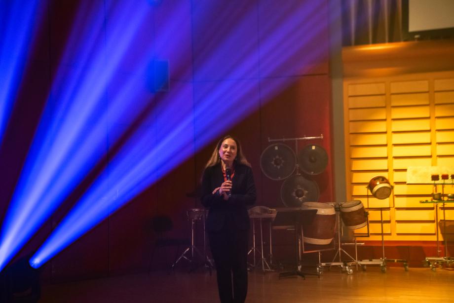 Polina Jakovleva (Leitung und Organisation des Auftritt der SuS des Musikgymnasiums) spricht mit Mikrofon auf der Bühne des Konzertsaals. Blaues Scheinwerferlicht scheint links von ihr, hinter ihr sieht man Schlaginstrumente.