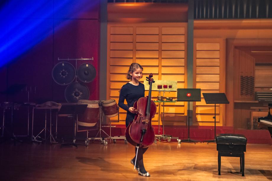 Die junge Musikerin Alexandra Wand ist mit ihrem Cello in Bewegung laufend auf der Bühne zu sehen. Hinter ihr sind Schlagwerk und einige Notenständer zu sehen.