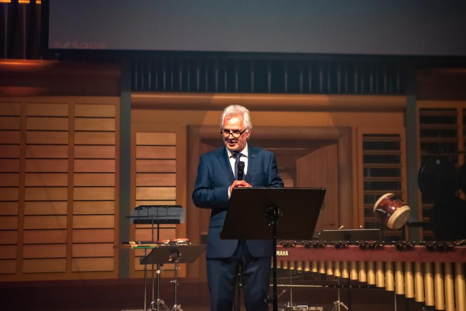 Rektor Axel Köhler bei seiner Eröffnungsrede. Er steht mit Mikrofon hinter einem Notenpult auf einer Bühne mit Schlaginstrumenten im Hintergrund