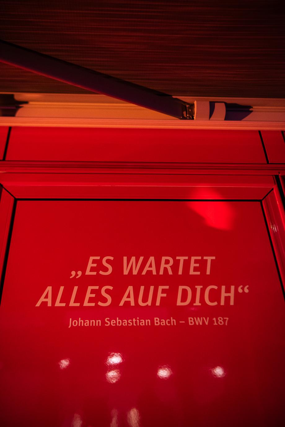 Nahaufnahme einer roten Fahrzeugoberfläche mit der weißen Aufschrift „Es wartet alles auf dich“ sowie dem Hinweis „Johann Sebastian Bach – BWV 187“, beleuchtet bei Nacht.
