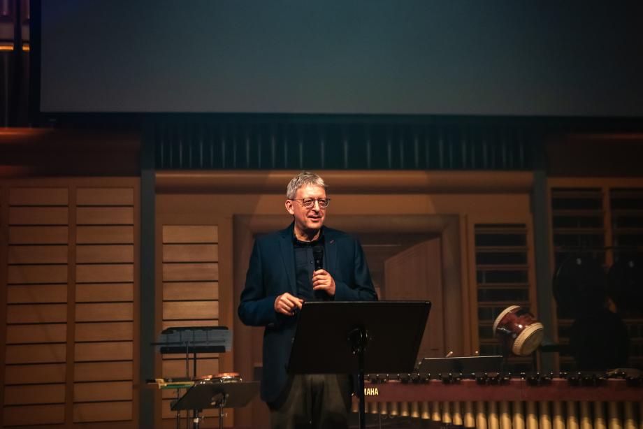 Hans-Christoph Rademann (Akademieleiter Internationale Bachakademie Stuttgart) steht mit Mikrofon auf der Bühne sprechend hinter einem Notenpult. Hinter ihm ist Schlagwerk zu sehen.