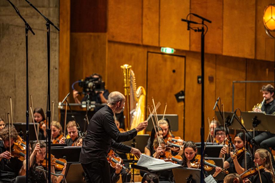 Im Zentrum steht der Dirigent Rasmus Baumann, der den Blickkontakt zu einer Instrumentengruppe sucht. Er ist umgeben vom Streicherapparat des Hochschulsinfonieorchesters.